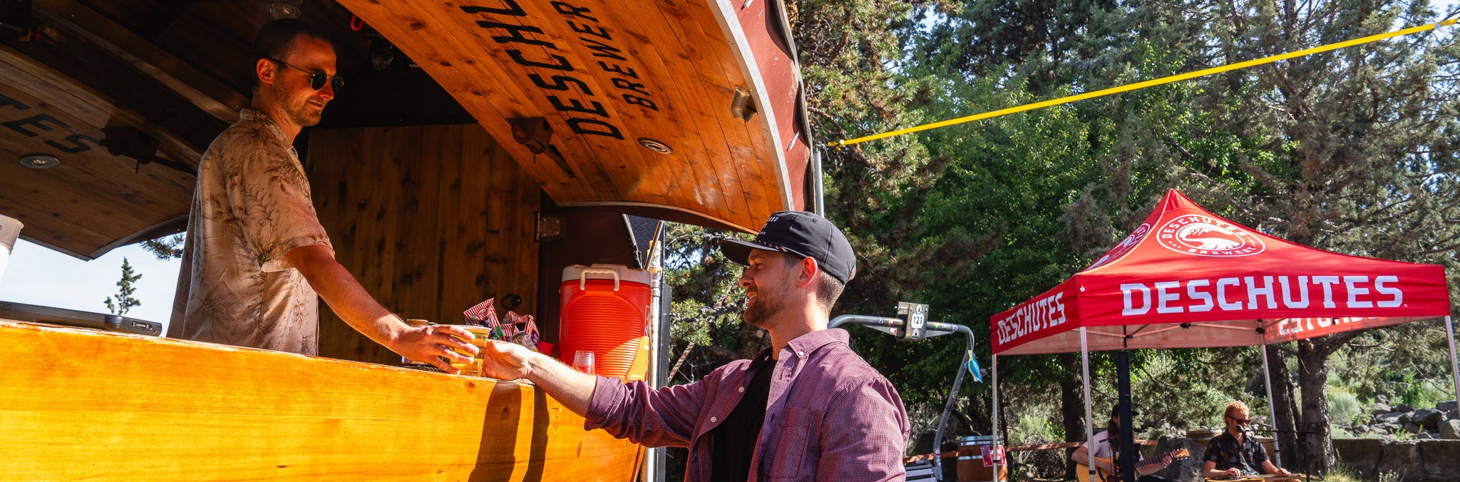 Two men interacting near a wooden truck with 'Deschutes' branding, set against a natural backdrop with trees and a red tent.