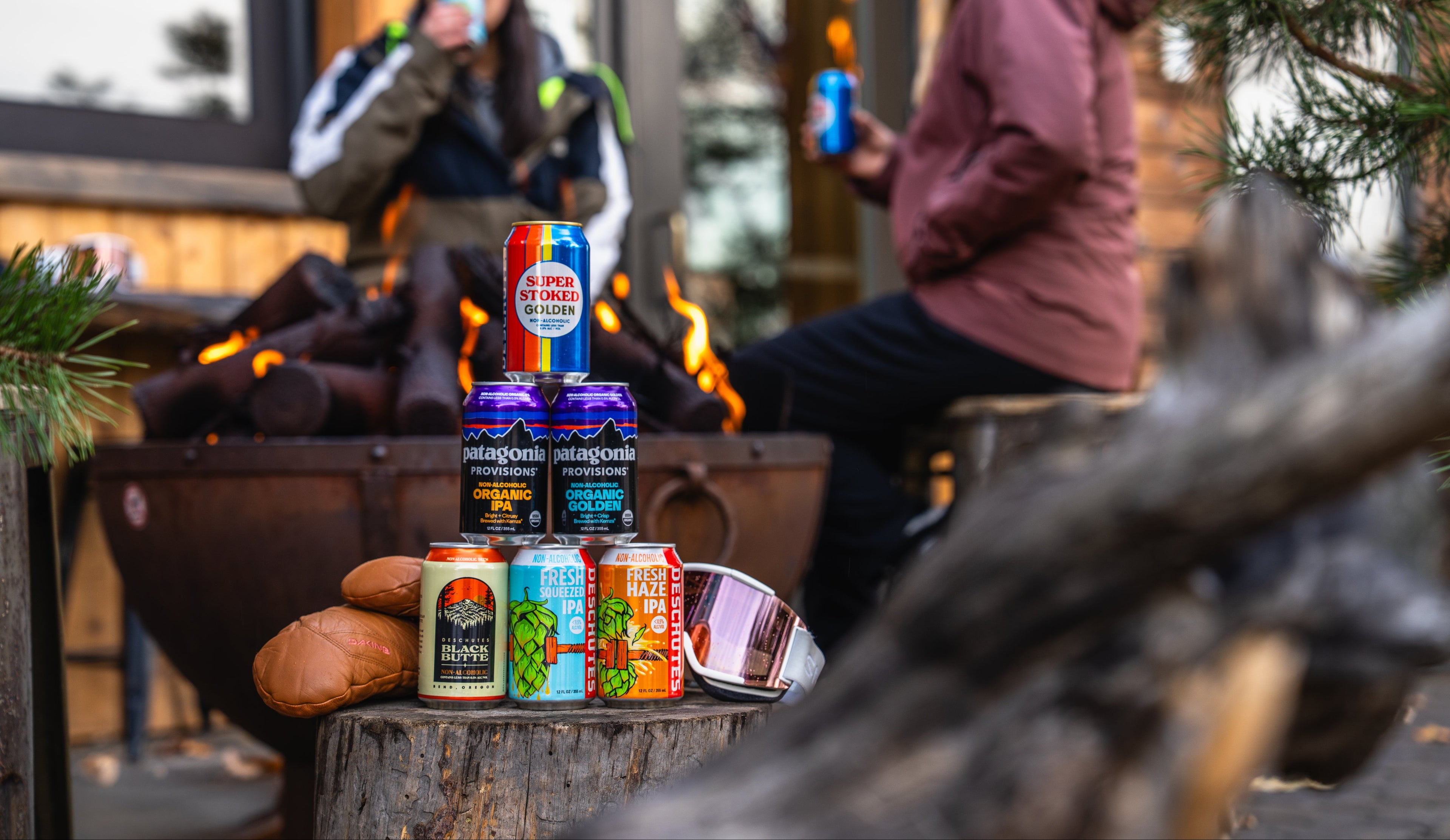Assorted cans of beer on a log with people by a fire in the background