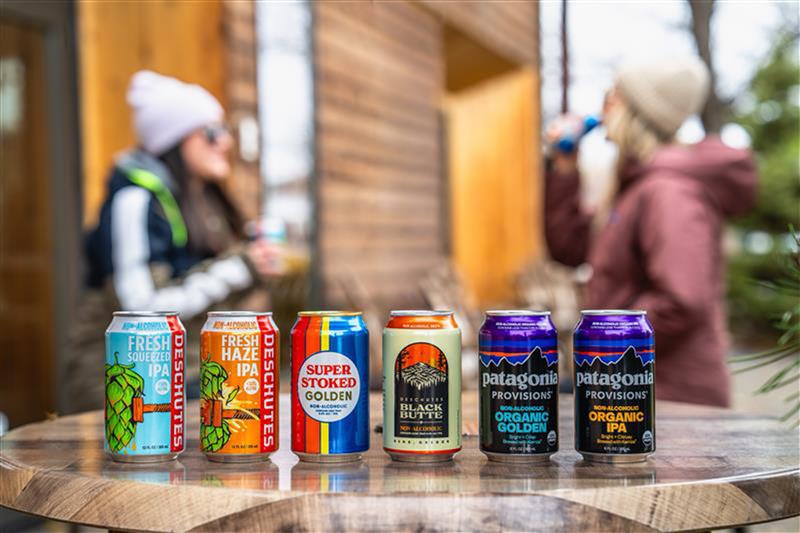 Row of Deschutes Non-Alcoholic beer cans on a wooden table with people sipping on them in the background