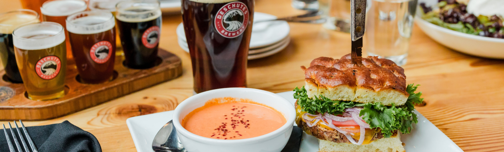 Burger and beer glasses on a wooden table with a bowl of soup.