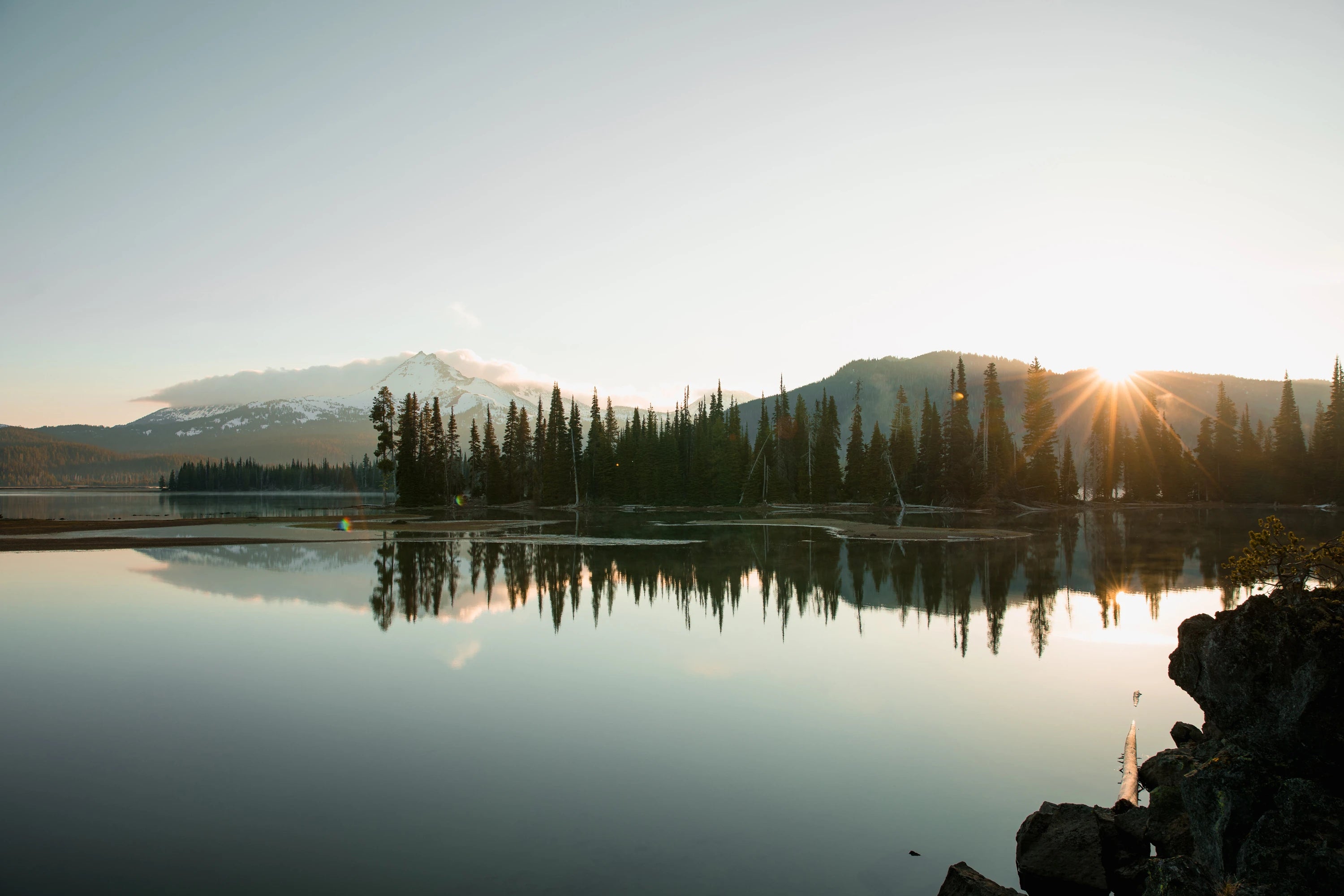 A photograph of a tree line beyond a lake, with mountains in the background.